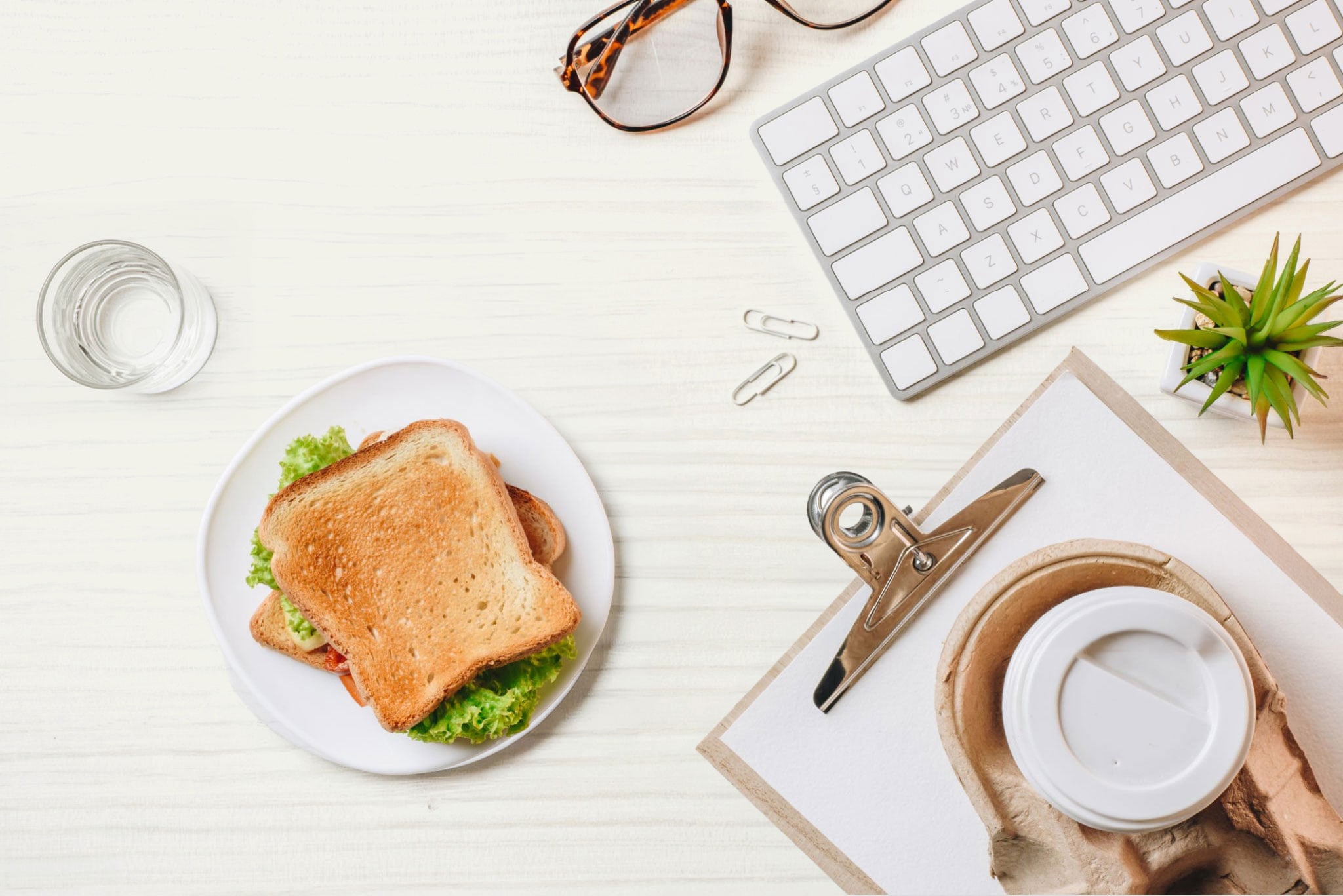 Overhead desk scene with sandwich, keyboard, coffee, and clipboard